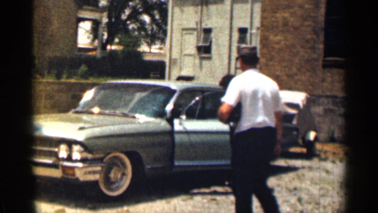 1963 Texas home-movie frame showing a man by an open pale green Cadillac door outside a brick residential building.
