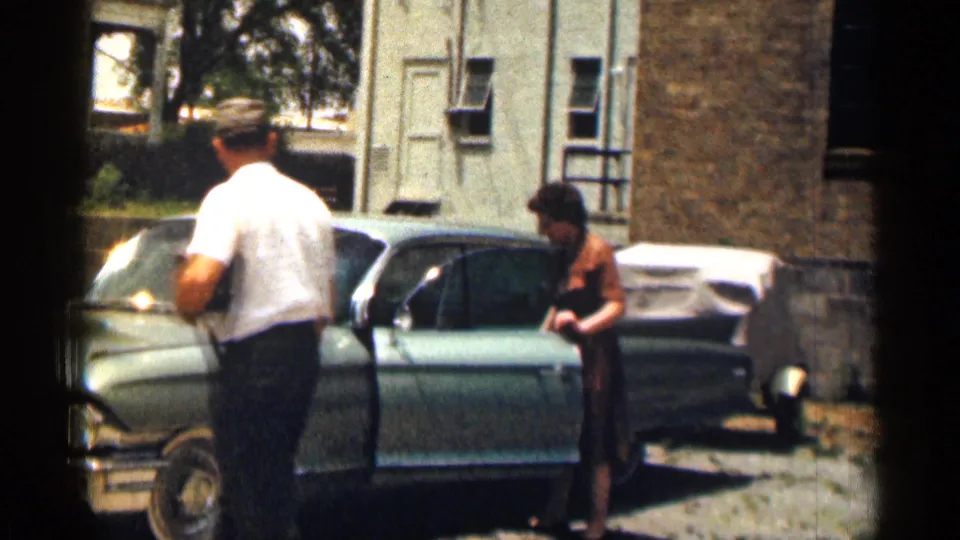 1963 Texas home-movie frame with a woman in a brown dress emerging beside a pale green Cadillac as a man stands nearby.
