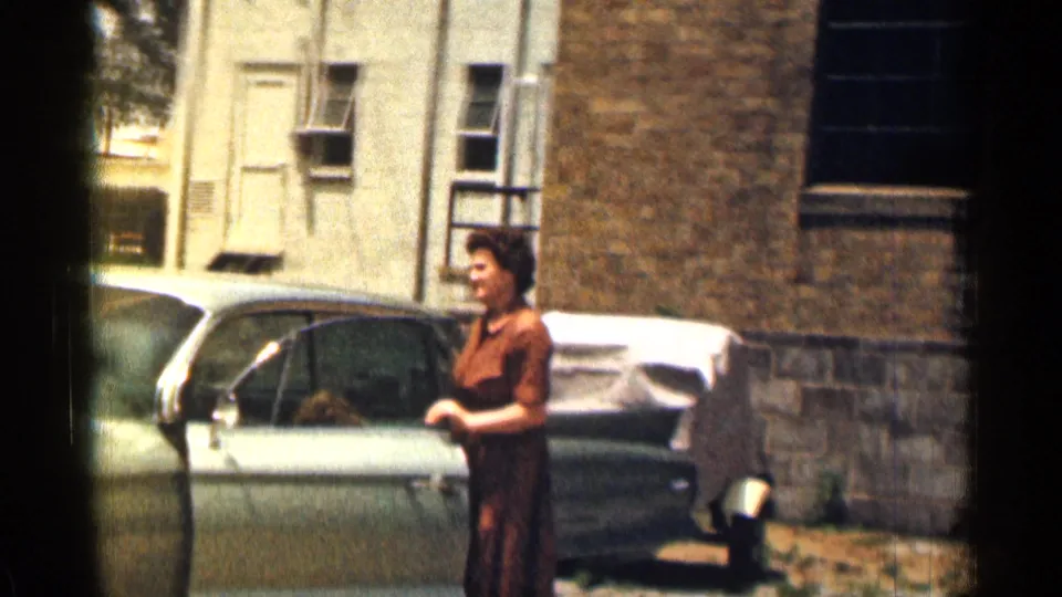 1963 Texas home-movie frame showing a woman with curled hair standing beside the open door of a pale green Cadillac.