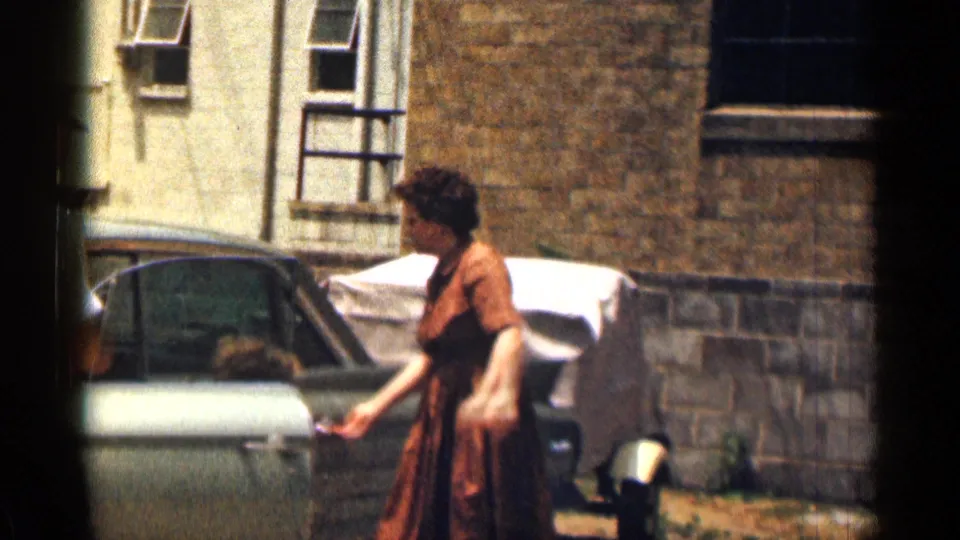 1963 Texas home-movie frame of a woman in a brown dress holding the open Cadillac door beside a brick exterior.