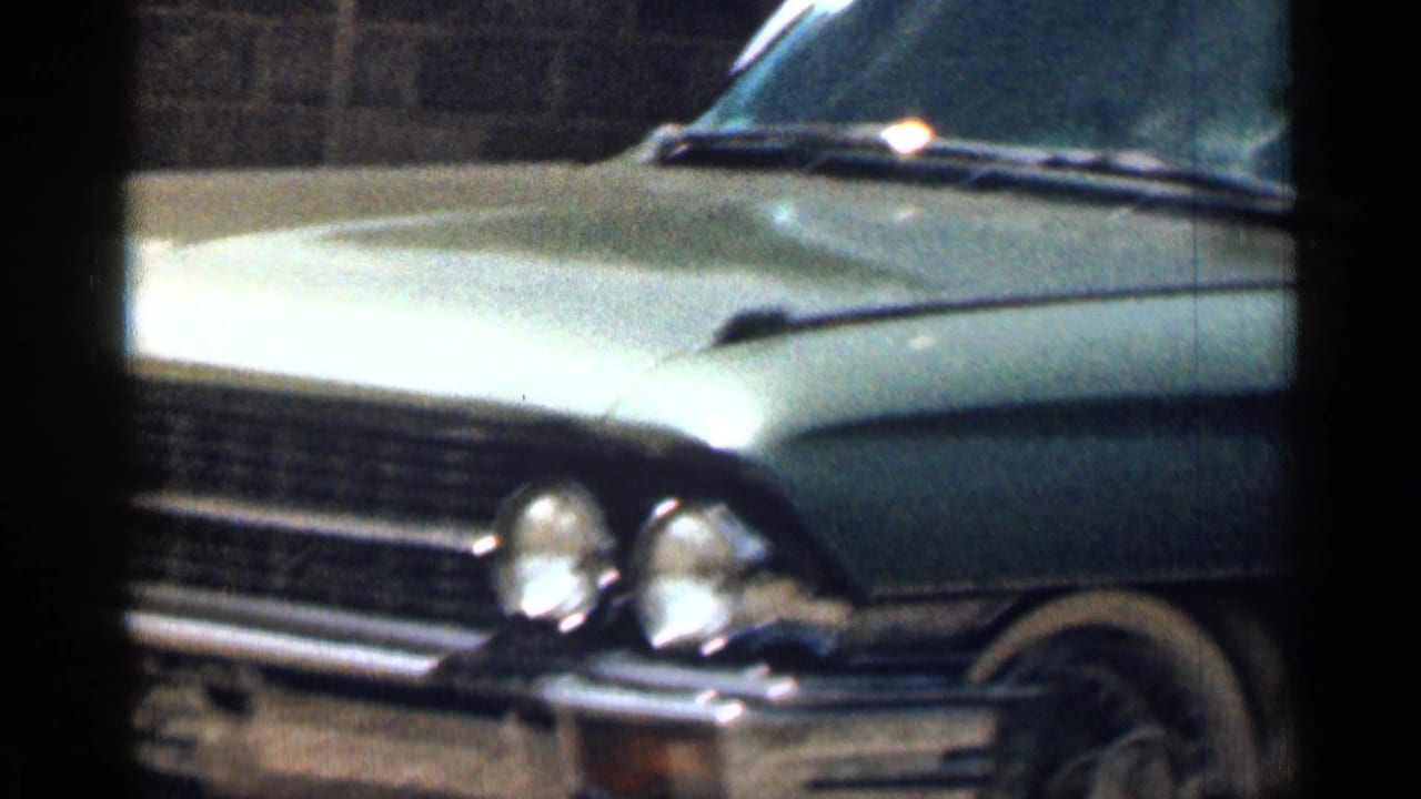 1963 8mm frame showing the front side of a light-colored vintage car with chrome trim in partial shade.