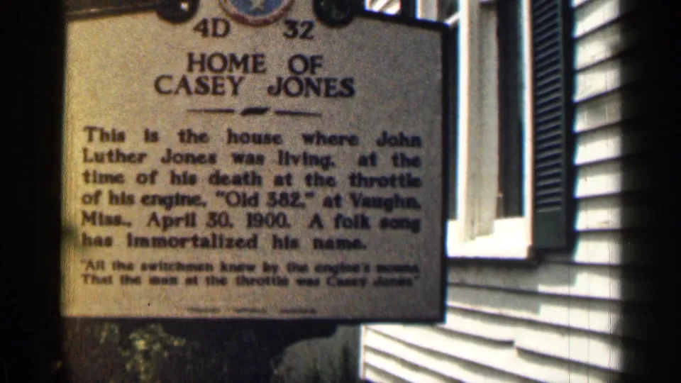 Final close frame of the Home of Casey Jones marker beside the white-sided building, with soft 8mm grain.