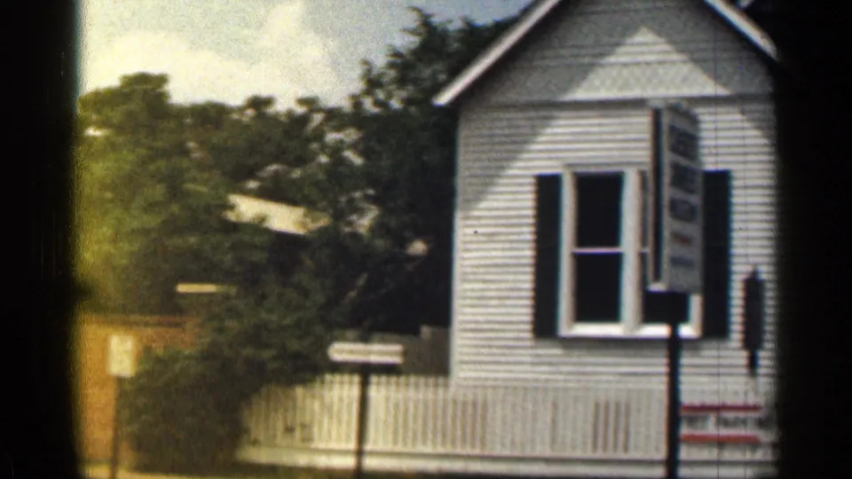 1963 Texas residential street frame with a white house, window sign, picket fence, trees, and small roadside signs.
