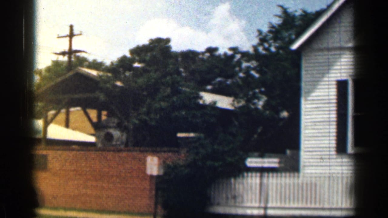 1963 Texas 8mm frame showing a white clapboard building, brick wall, trees, utility pole, and pale sky.