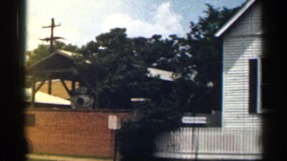 1963 Texas 8mm frame showing a white clapboard building, brick wall, trees, utility pole, and pale sky.
