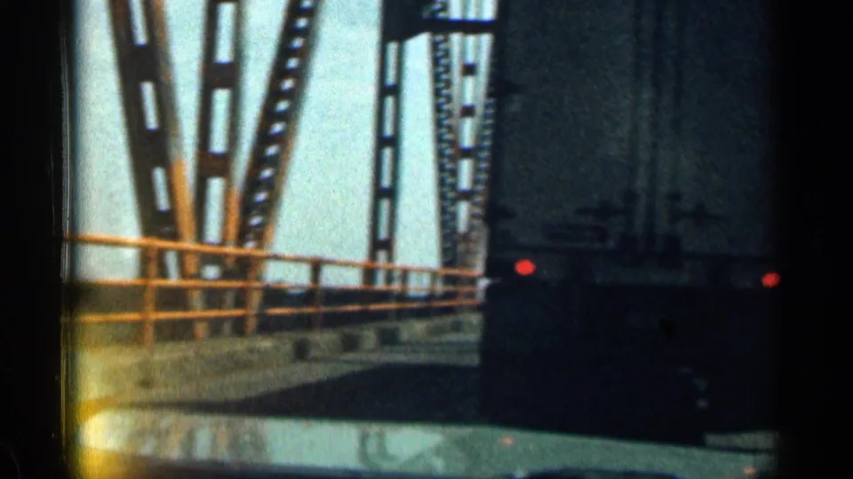Passenger-view 8mm frame from a moving vehicle crossing a metal truss bridge with railings and a pale sky.