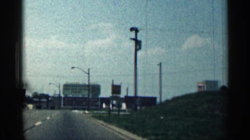 1963 Texas roadside scene with a paved road, utility poles, large highway signs, and a grassy embankment from a moving vehicle.