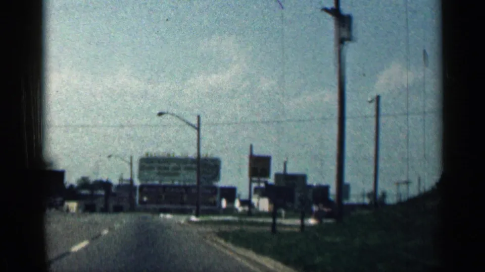 1963 Texas home-movie view from a moving vehicle approaching roadside signage, streetlights, and low commercial buildings.