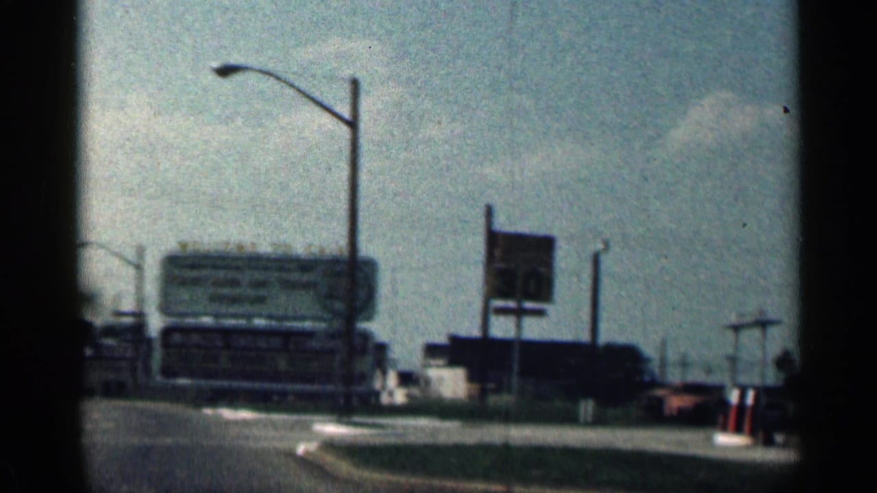 Road curves past large roadside signs, streetlights, and a red car at the right edge.