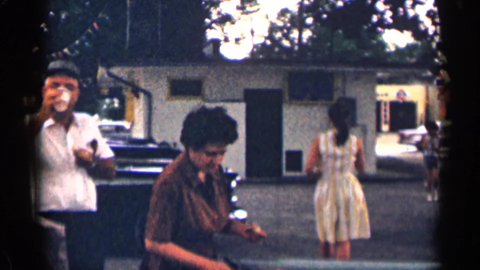 Casual 1963 Texas roadside stop with a man drinking from a cup, another person looking down, and a woman walking near parked cars.