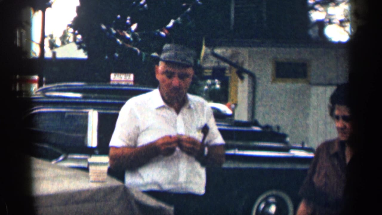 1963 Texas home-movie frame of a man in a white short-sleeve shirt and cap standing near a parked car, with a woman partly visible nearby.