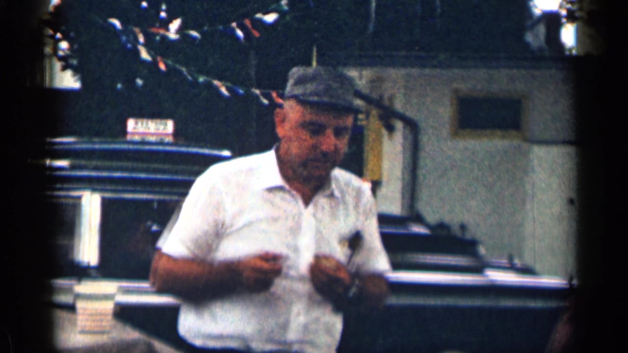 Man in a cap stands in front of a dark parked car, holding a small object near his shirt.