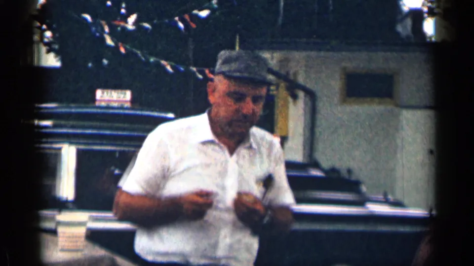 1963 Texas 8mm frame showing a man in a white shirt and cap near a dark parked car and building, with string lights overhead.