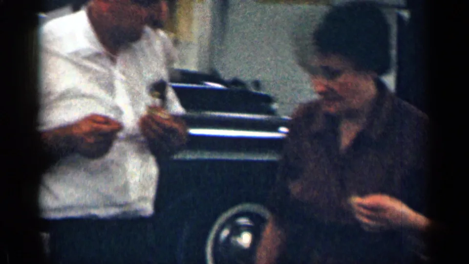 1963 Texas home-movie frame of a man and woman standing near a vintage car, holding small food items or objects in a parking lot.