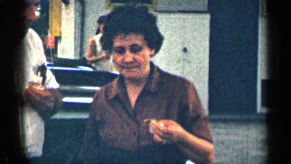 1963 Texas 8mm frame of a woman in a brown blouse holding food in a parking lot, with parked cars and a building behind her.