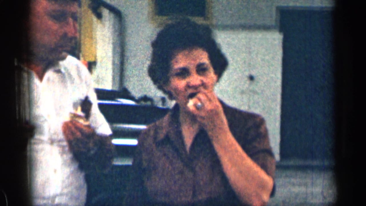 Older man and woman eating sandwiches outdoors beside a parked car and building.