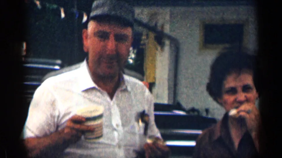An older man in a white shirt holds a striped coffee cup and food as the woman eats slightly behind him in the 1963 Texas scene.