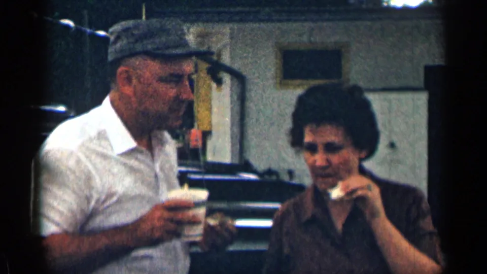 The couple appears to converse while holding a coffee cup and sandwiches, framed by a parked car and building exterior in Texas.
