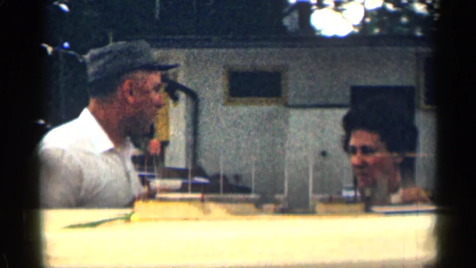 In 1963 Texas 8mm footage, a man in a white shirt and cap talks with a woman beside lodge windows and walls.