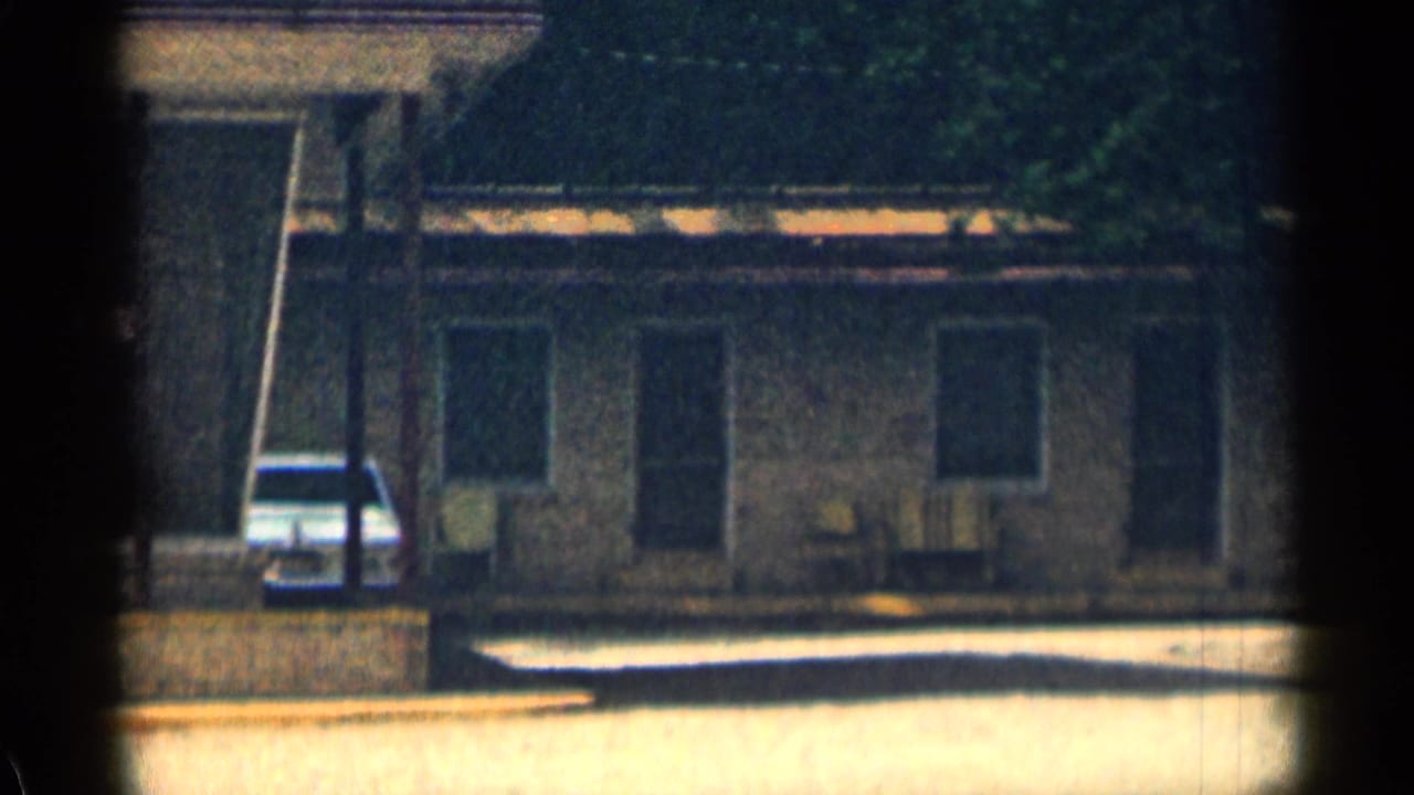 Motel building exterior with windows, chairs, trees, and a parked white car.