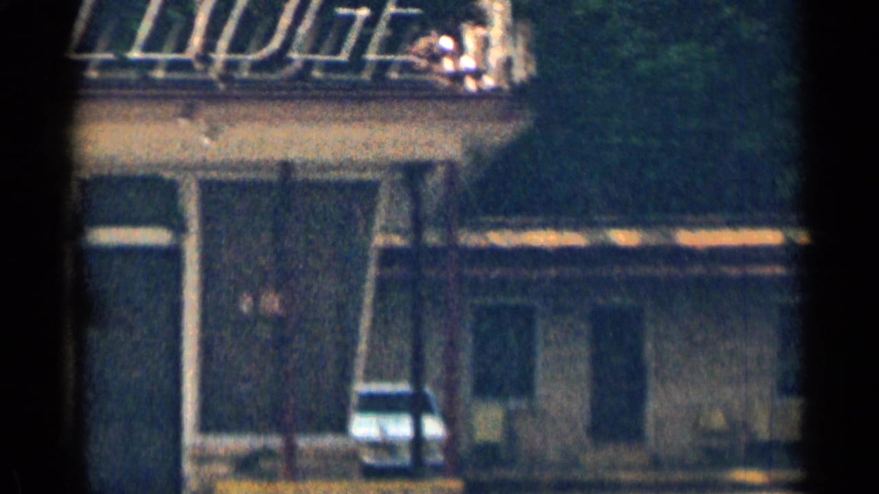 Motor lodge exterior with balcony railing, doorway, parked car, and chairs.