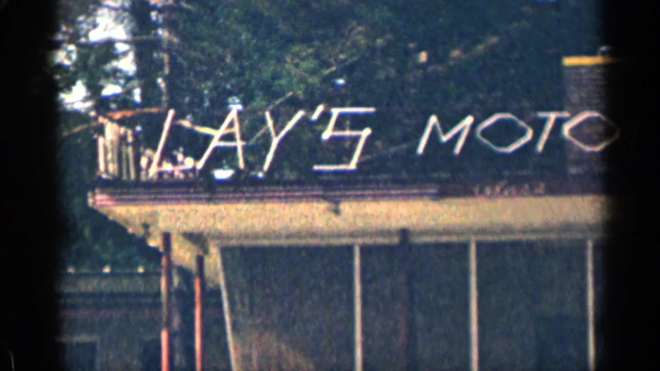 1963 Texas home-movie frame showing a Lay's Motor sign across a low building roof with trees behind it.