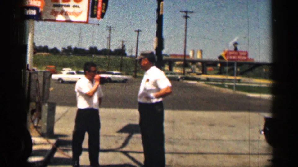1963 Texas 8mm frame of two adult men standing near gas pumps and roadside signs under a bright blue sky.