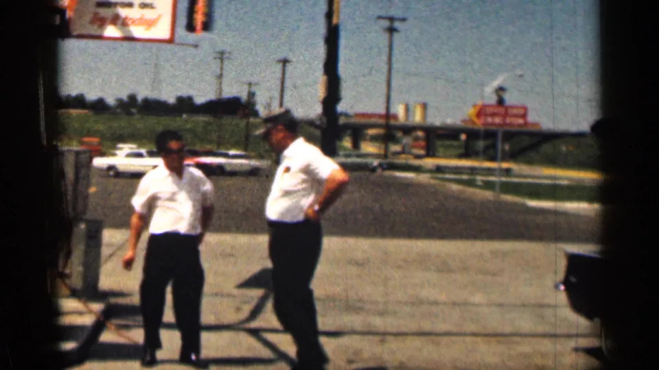 1963 Texas home-movie frame showing two men on a gas station forecourt, with cars, utility poles, and signs in the background.