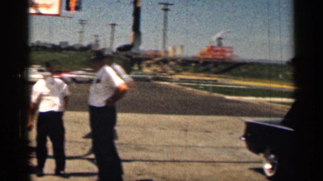 1963 Texas home-movie frame showing two men in white shirts near a gas station area with pavement, signs, and a partial car visible.