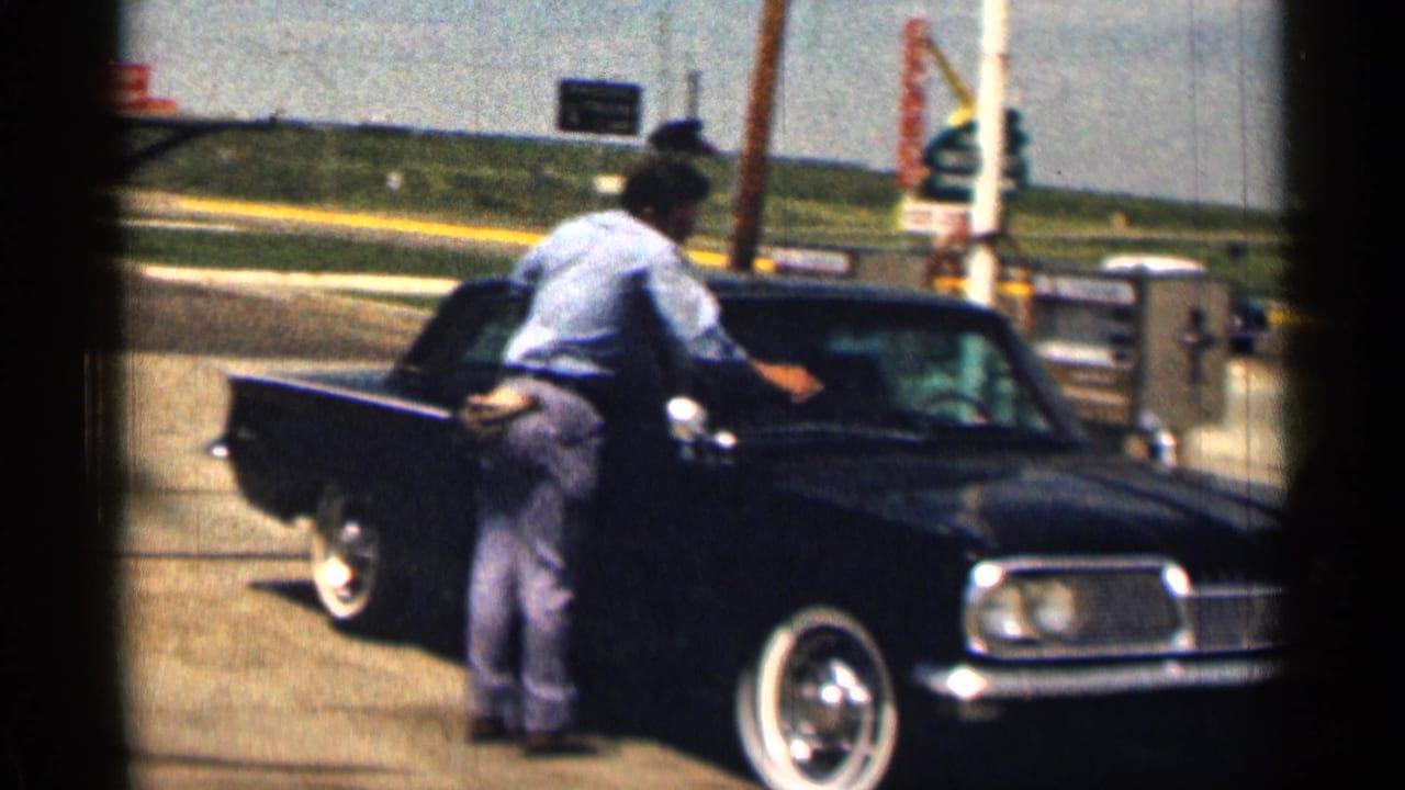 A man leans into a dark classic sedan beside a gas pump area.