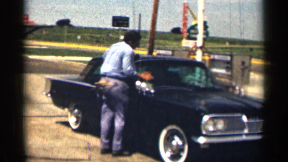 1963 Texas home-movie still with a man beside a dark classic sedan, whitewall tires, pump equipment, and roadside signage.