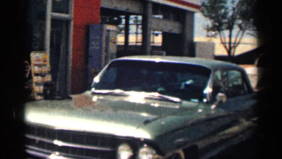 1963 Texas frame showing a light-colored classic sedan parked near a roadside commercial building with windows and signage.