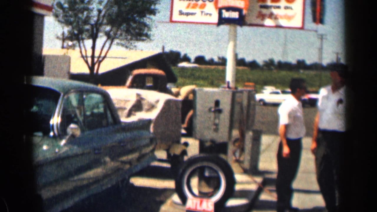 Green sedan beside service equipment with two standing figures near the forecourt.
