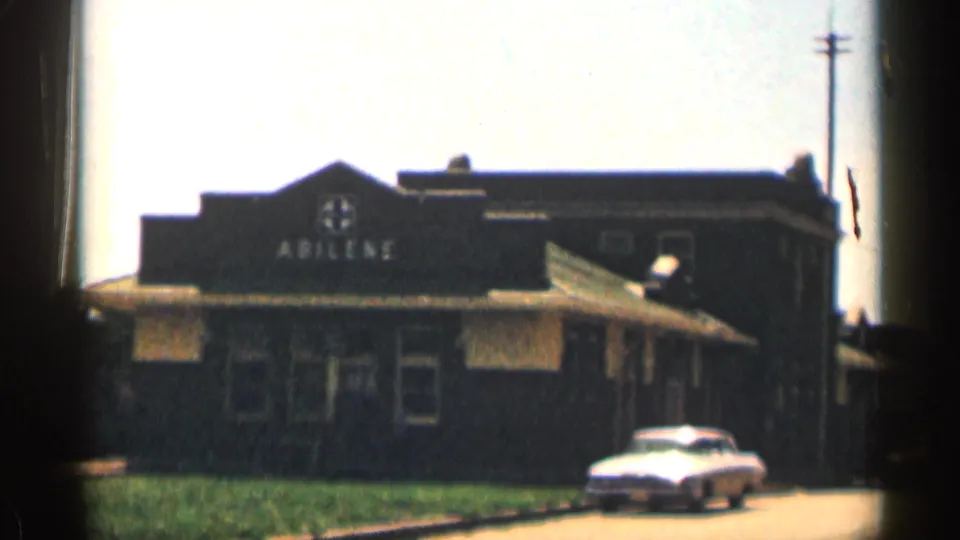 Wider 1963 Texas 8mm view of the Abilene-labeled building, utility pole, grass, sidewalk, and white sedan.
