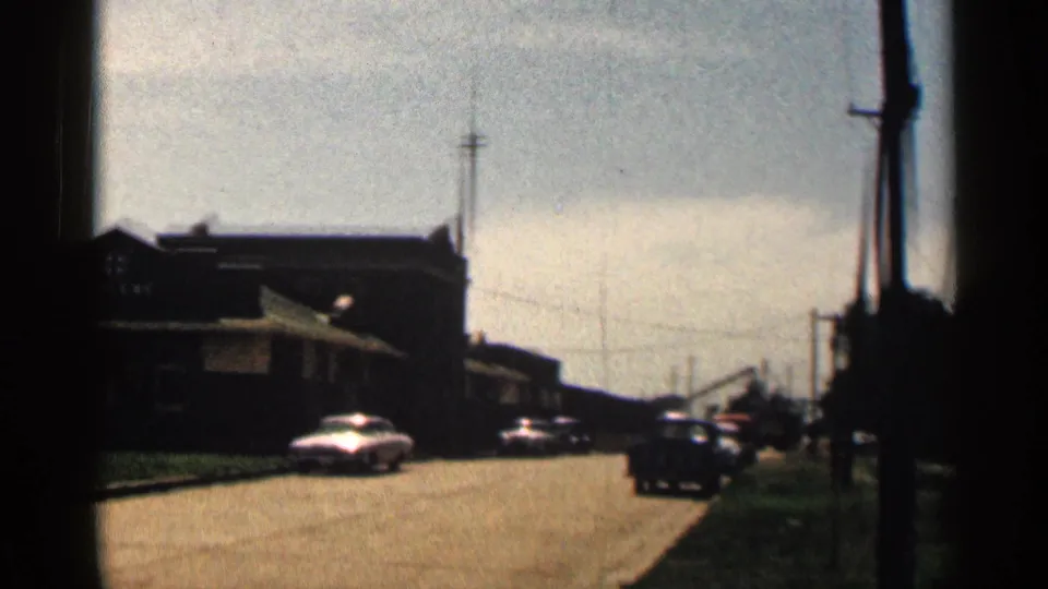 1963 Texas street view with a dark building, classic cars, utility poles, and a hazy sky during a handheld pan.