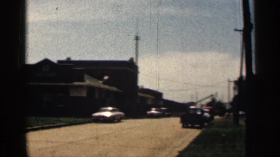 1963 Texas frame showing classic cars on a broad road beside a low dark building, grass verge, poles, and soft sky.
