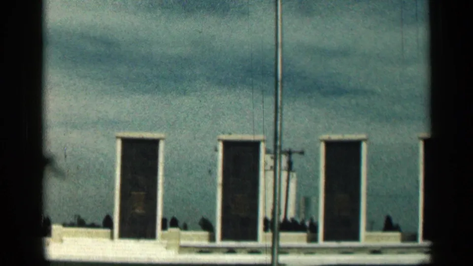 1963 Texas 8mm frame of a blue building facade with narrow dark doors, white trim, utility lines, and a greenish sky cast.