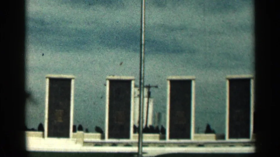 1963 Texas 8mm frame showing a flagpole partly blocking a low memorial wall with dark rectangular plaques and pale trim.