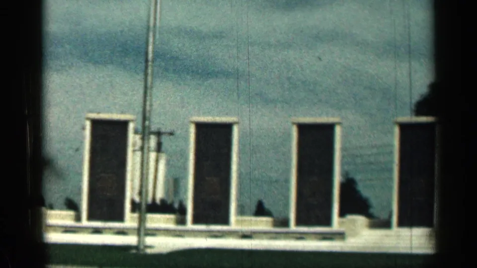 1963 Texas 8mm view of dark memorial plaques on a pale low wall, crossed by a tall flagpole and bordered by trees.