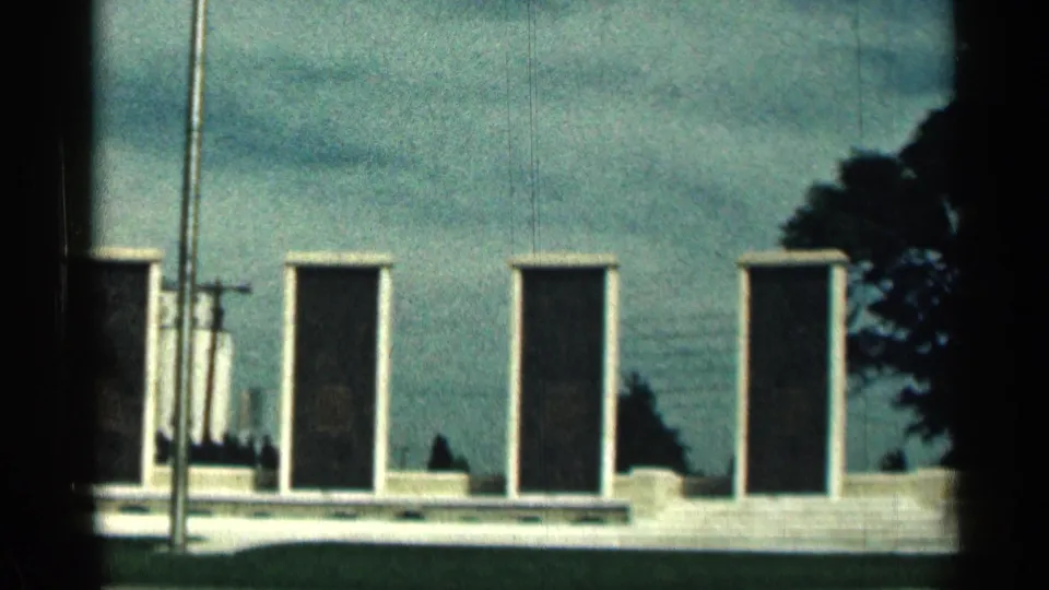 1963 Texas home-movie frame with the memorial wall cropped closer, showing plaques, flagpole hardware, shrubs, and cloudy sky.