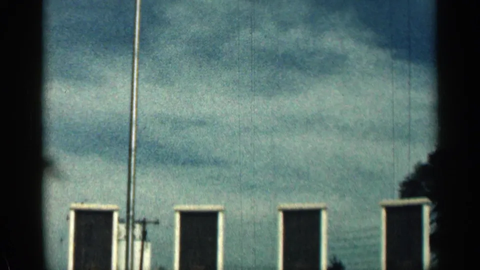 1963 Texas 8mm frame emphasizing the tall flagpole, cloudy sky, and spaced dark plaques along the memorial wall.