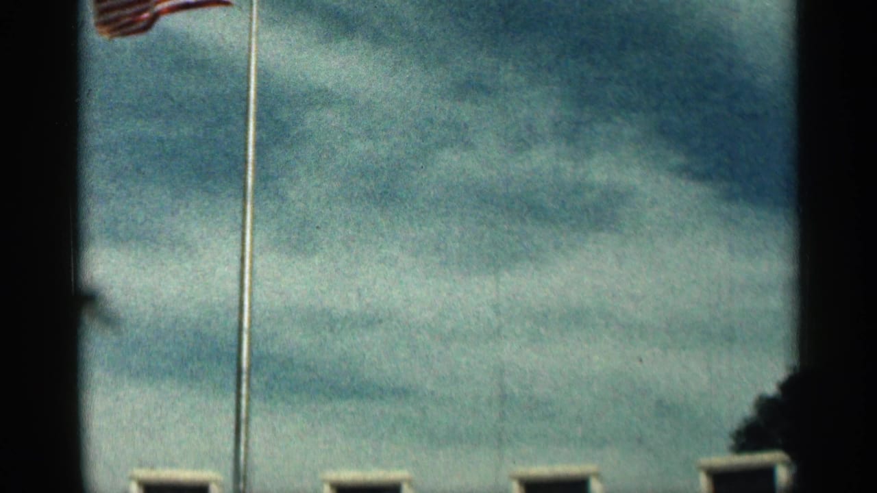 1963 Texas 8mm frame showing an American flag on a flagpole against cloudy blue sky, with dark gate edges and soft exposure.