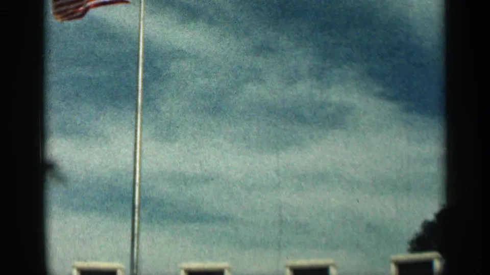 1963 Texas 8mm frame showing an American flag on a flagpole against cloudy blue sky, with dark gate edges and soft exposure.