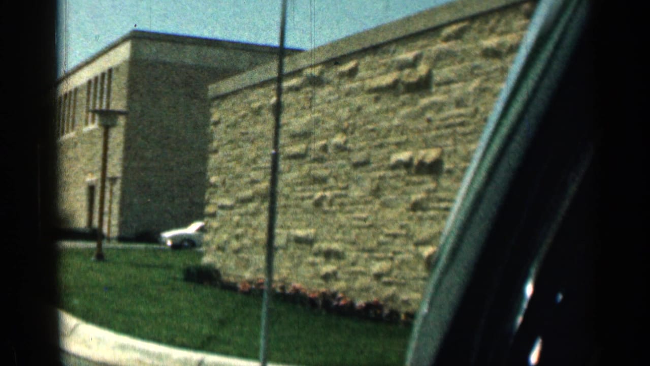 Brown stone building, green lawn, white car, and curb seen from a vehicle window.