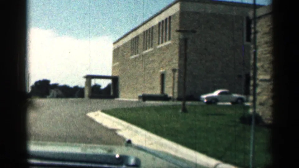 1963 Texas 8mm frame of a low brown stone building with upper windows, lawn, curb, small entry structure, and a parked white car.