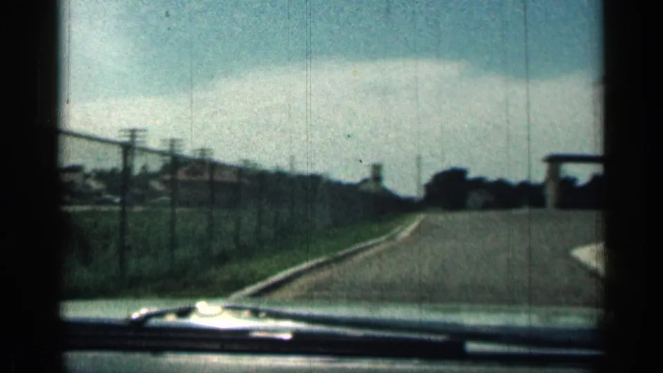 1963 Texas home-movie view down a road beside a chain-link fence, with windshield edge visible and trees in the distance.