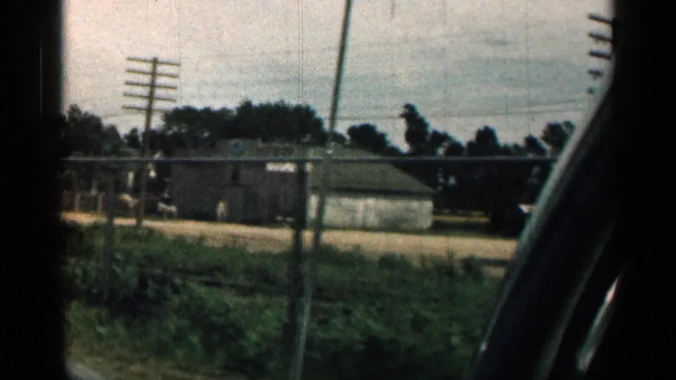 Passenger view from a moving car in 1963 Texas, with low roadside buildings, utility poles, fencing, trees, and a dark interior window frame.
