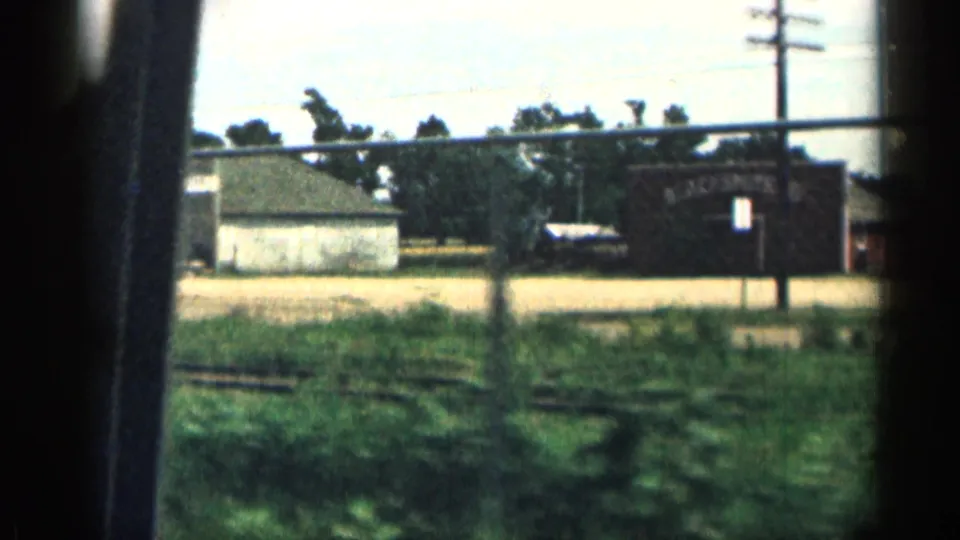1963 Texas car-window view of a fenced roadside area with low buildings, grass, utility poles, trees, and a dark building with a bright doorway.