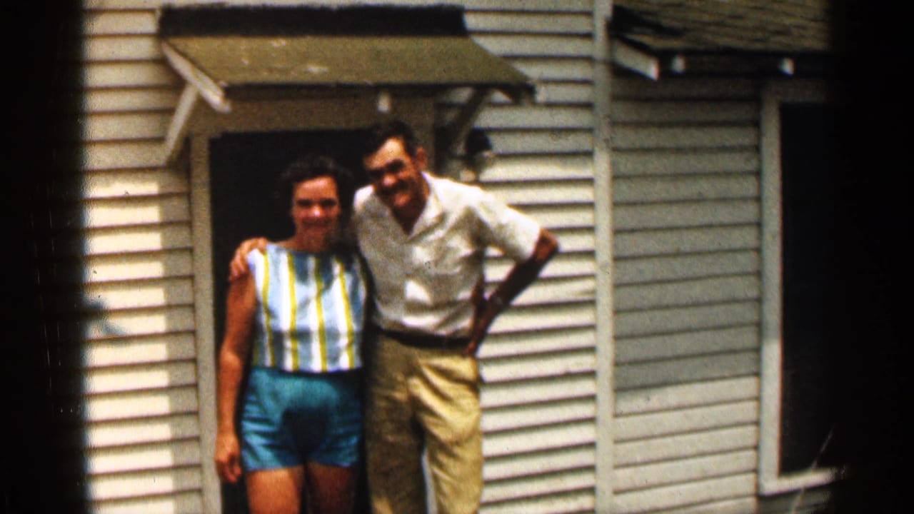 A smiling woman and man stand arm in arm by a white-sided house.