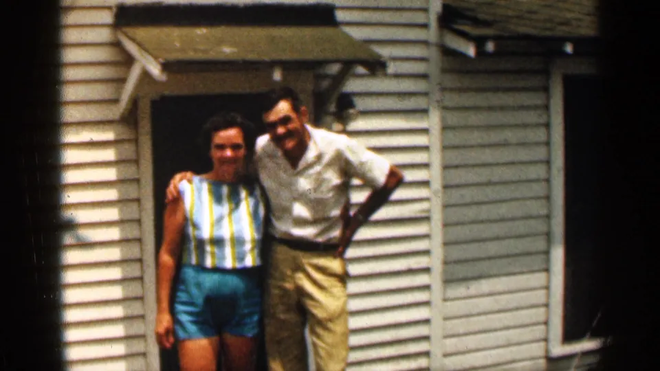In this 1963 Texas frame, a smiling couple stands close together outside a white-sided house with a doorway awning.